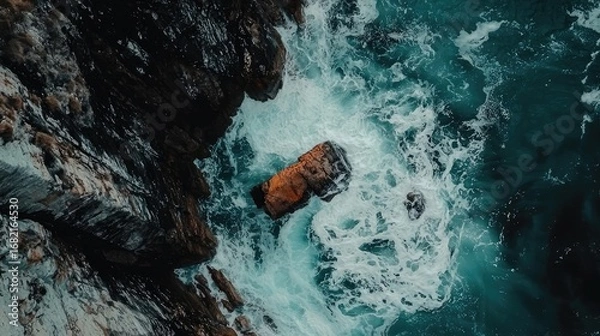 Fototapeta Aerial View of Ocean Waves Crashing Against Dark Rocks Along the Coastline with Turquoise Water and White Foam Creating a Dynamic Seascape