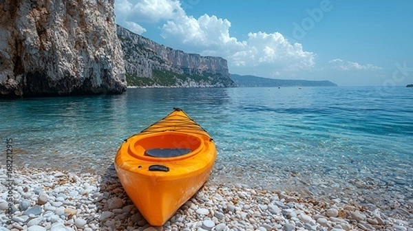 Fototapeta Orange kayak on pebble beach near calm turquoise sea and cliffs.