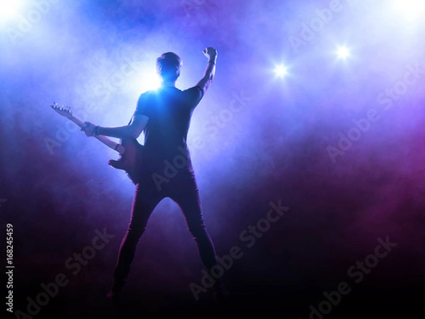Obraz Silhouette of guitar player on stage on blue background with smoke and spotlights	