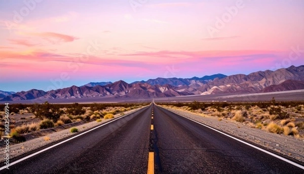 Fototapeta A straight asphalt road runs to the horizon, flanked by desert scrub and distant mountains under a vibrant, colorful sunset sky
