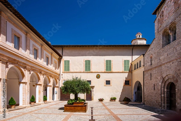 Fototapeta Wide angle view capturing the cloister of Spoleto Cathedral, featuring elegant arches, a flourishing olive tree, and a vibrant blue sky on a sunny summer day in Umbria, Italy