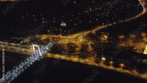 Fototapeta Aerial shot of glowing Budapest with bright bridge across dark river in night time, Hungary