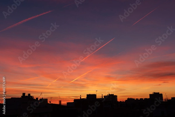 Fototapeta A vibrant sunset with shades of orange, pink, and purple fills the sky. Silhouettes of buildings and contrails are visible against the colorful backdrop.