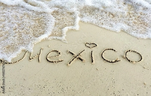 Fototapeta The inscription on the sand on the beach "Mexico" and the foam waves of the sea. Travel background with sign text on sandy coast. Vacation on Caribbean