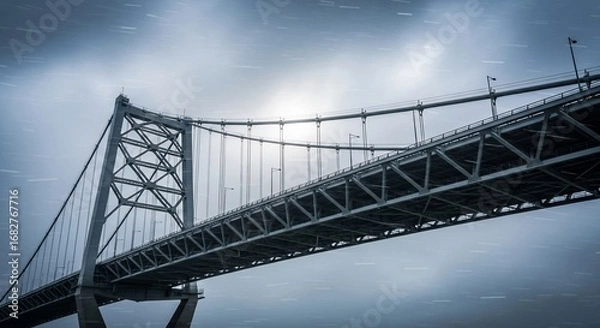 Obraz A low-angle perspective of a massive steel suspension bridge under a dramatic and gloomy overcast sky.