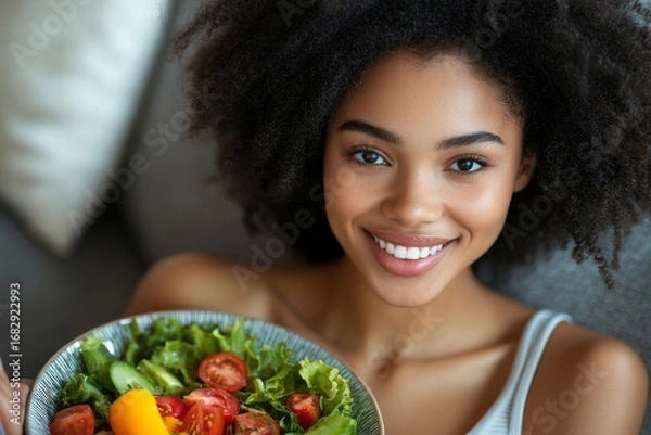 Fototapeta Portrait of a young woman sitting at home on the sofa with a laptop, eating salad and fresh vegetables from a plate, smiling at the camera, Generative AI
