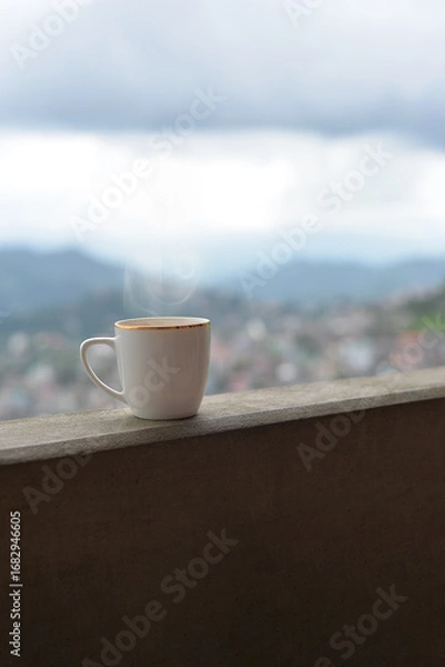 Fototapeta A steaming cup of tea rests on a concrete balcony, with blurred hills fading softly in the background
