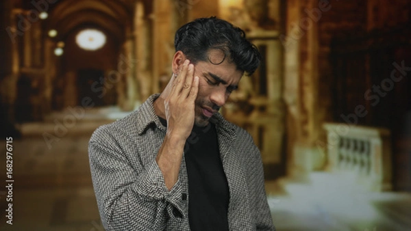 Obraz Young man in grey jacket holding cheek inside ornate church building; toothache discomfort concern worry.