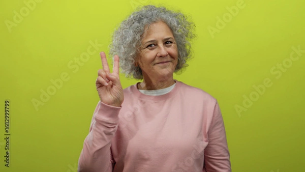 Obraz Woman with grey hair smiling and showing peace sign against a vibrant yellow background wearing a pink sweater conveying happiness and positivity.