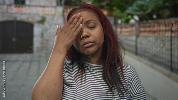 Obraz Woman touches forehead wearing wooden watch on cobblestone street outside brick building lined with greenery; stress.