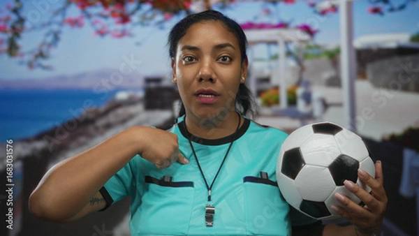 Obraz Woman referee in turquoise jersey holds soccer ball on seaside promenade while pointing finger to chest; confidence.