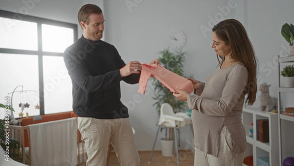Obraz Pregnant woman and man prepare nursery in baby room with crib, displaying new pink baby clothes in a cozy home setting, emphasizing family bonding and anticipation.