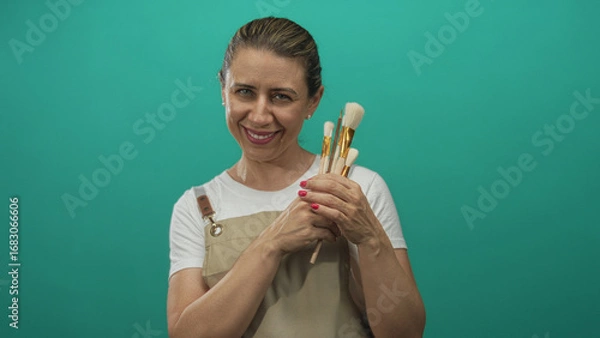 Obraz Woman holding paintbrushes wearing a beige apron and smiling in a studio with teal walls; creativity joy.