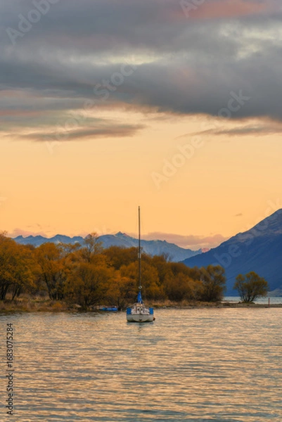 Fototapeta Scenic view of sailboat floating on Lake Wakatipu surrounded by golden autumn trees and mountain landscape under sunset sky in New Zealand