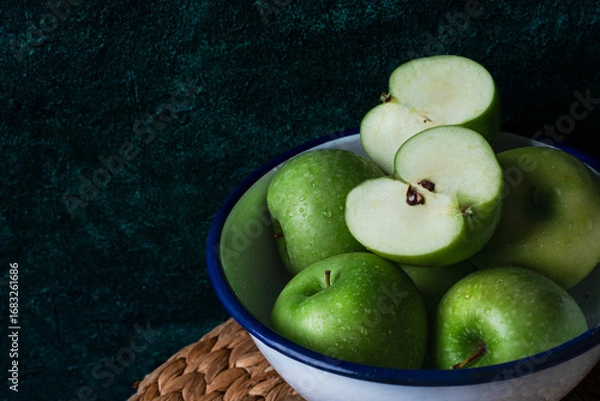 Fototapeta Close-up of fresh green apples in a white enamel bowl with blue rim, placed on a rustic mat against a dark textured background.