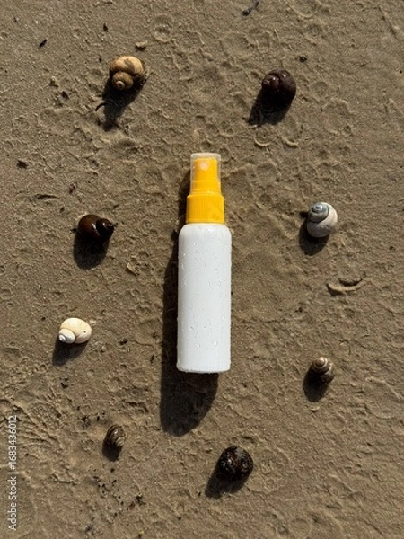 Obraz A white plastic spray bottle with a yellow cap placed on wet sand, surrounded by seashells. Minimalist beach-themed composition, suitable for concepts of summer, skincare, sunscreen, vacation