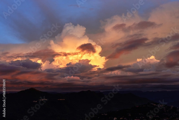 Fototapeta Golden sunset clouds blaze through the evening sky, casting a warm glow over the dark, quiet mountain silhouettes below.