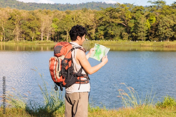 Fototapeta An Asian young man traveler looking at an old paper map to find a route beside a lake during sunny day