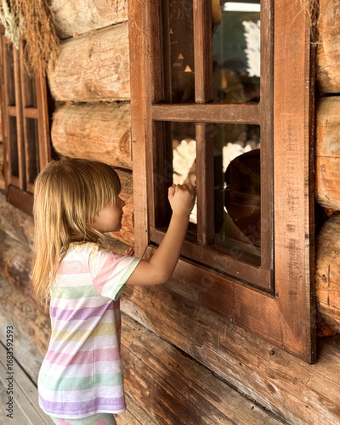 Fototapeta A child looks into the window of a wooden house.