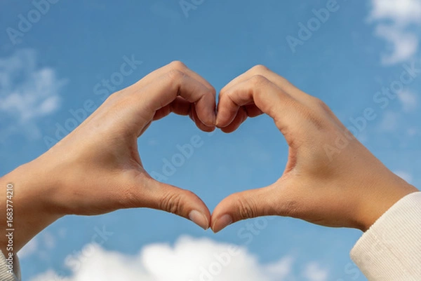 Fototapeta Two hands forming a heart shape against a blue sky with clouds love symbol