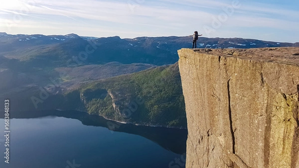 Obraz A hiker stands with arms outstretched in a gesture of freedom on the edge of Preikestolen, taking in the vast and epic panoramic view of the Lysefjord and distant snow-capped mountains in Norway.
