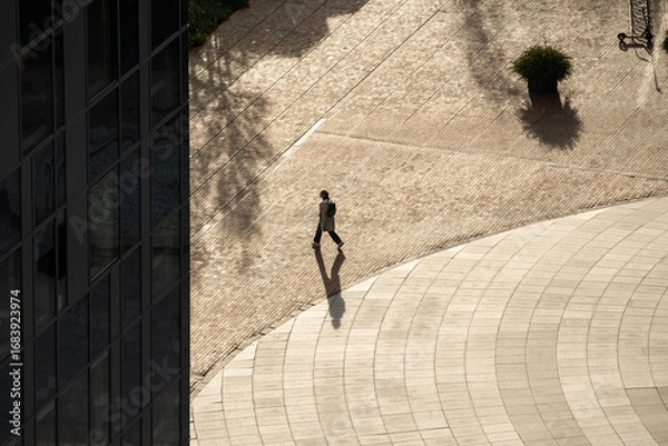 Fototapeta A solitary person walks through a sunlit urban plaza, casting a long shadow on the patterned ground. The scene captures architectural lines, open space, and minimalism in a city environment