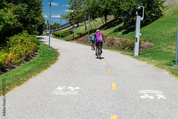Obraz Bike path surrounded by trees and modern infrastructure. Cyclists on the path.