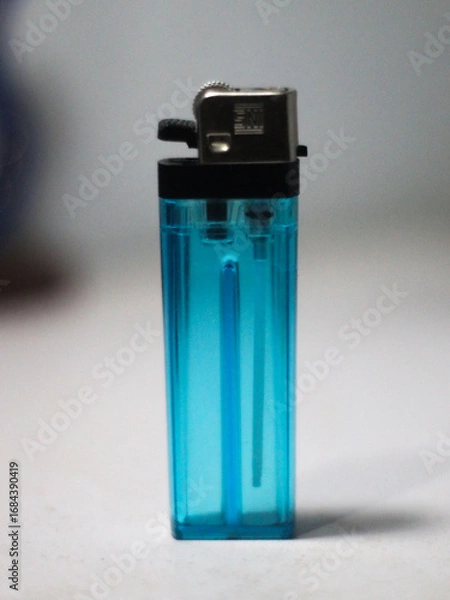 Obraz This photo shows a blue transparent gas lighter with a silver metal head. The lighter stands upright on a white surface, with its simple details clearly visible.