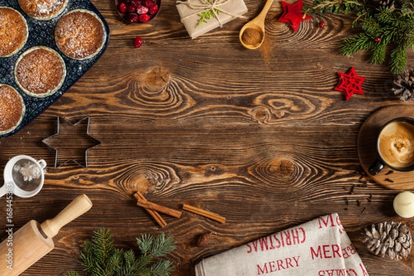 Fototapeta Christmas cookies with fir-tree branch on old wooden background. Cookies, black cup, candle, cupcakes, cone, cinnamon, wooden spoon, gift, branches of fir-tree. Copy space. Top view