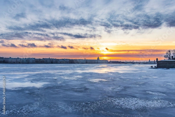 Fototapeta Dramatic winter sunset over a frozen river with colorful sky, clouds, and silhouettes of historical buildings on the horizon. The frozen surface reflects the light of the evening sun, creating a