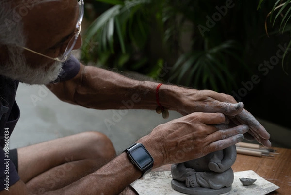 Obraz Indian old man crafting an eco-friendly clay Ganesh idol for Ganesh Chaturthi and teaching kids how to do it, sitting outdoors in a garden
