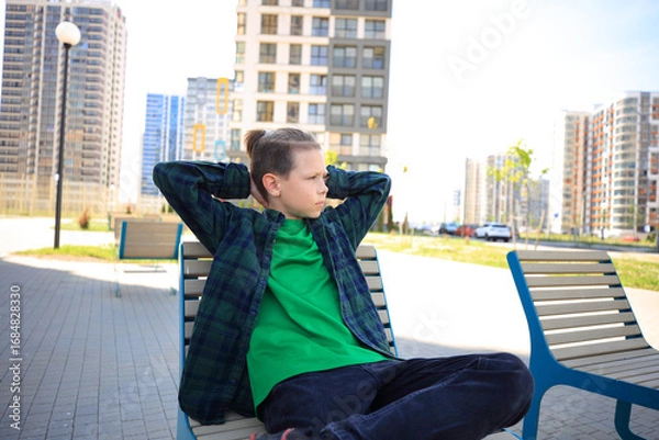 Fototapeta a teenager sitting alone on a bench, looking bored amidst the imposing of the city on a summer day. urban environment and the teenager's expression, isolation and longing for connection.