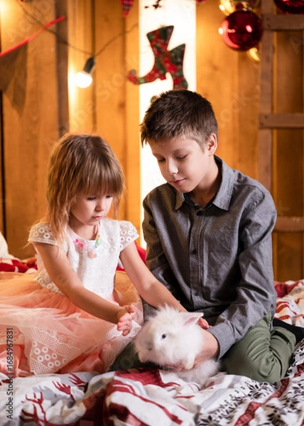 Fototapeta Boy and girl playing with white rabbit on bed. New Year mood. New Year decor.