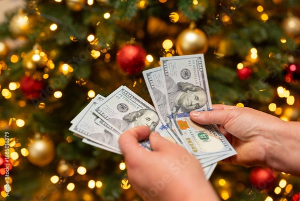 Obraz A person counting dollars against the background of a Christmas tree. Woman counting money near the Christmas tree, idea of big spending on gifts, decorations for New Year holidays