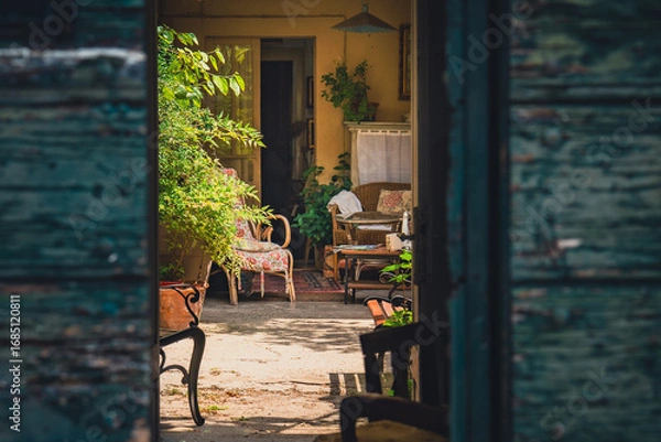 Fototapeta Rustic Italian patio with vintage furniture, plants and cozy atmosphere, captured through open wooden doors. Warm light, intimate mood, Mediterranean style.