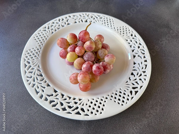 Obraz Decorative white carved plate holding a bunch of pink grapes on a dark surface, photographed from above, elegant and minimal still life composition.