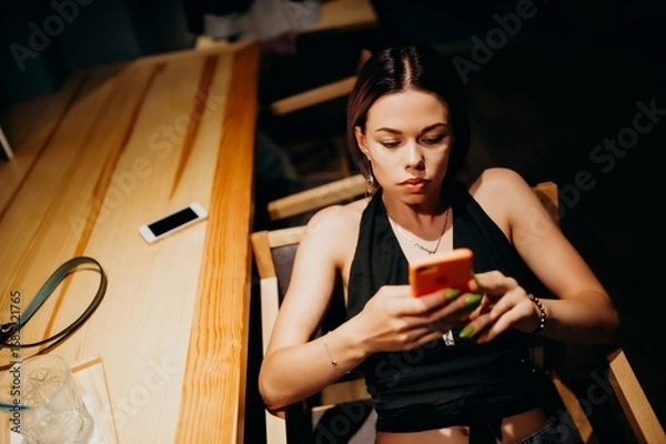 Fototapeta A young woman sits at a wooden table in a dimly lit room, focused on her phone. Dressed in a black top and jeans, she shows a calm yet concentrated mood, illuminated by warm light.