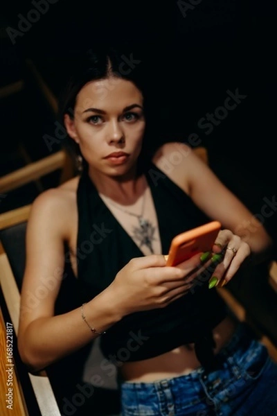 Fototapeta Close-up of a young woman with intense eyes, holding a phone. She is dressed in a black halter top and jeans, her green nails and jewelry adding a bold accent to the dark background.