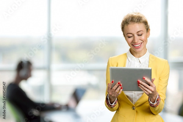 Obraz Smiling business lady with pc tablet. Cheerful young business woman looking on computer tablet and smiling, office window background.