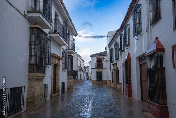 Fototapeta Ronda, Spain - A quiet cobblestone street glistens after spring rain. Whitewashed buildings and wrought iron balconies reflect the charm of this historic mountain town.