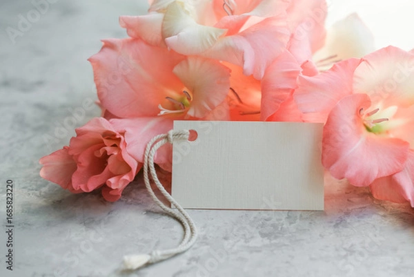 Fototapeta Blank gift tag rests among delicate pink gladiolus flowers on soft gray background. Ideal for mockups, invitations or social media posts