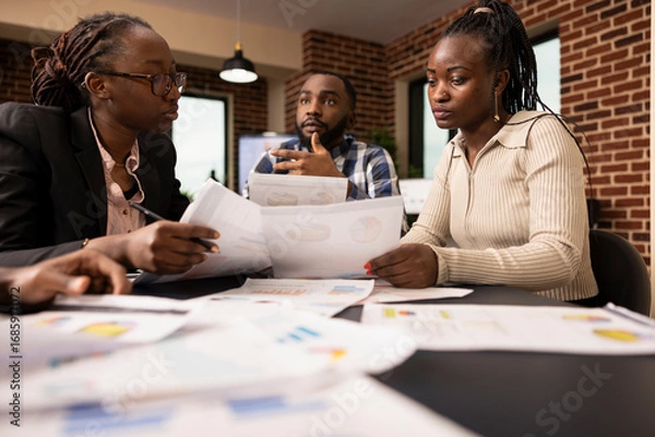 Fototapeta African american entrepreneurs with documents discussing and consulting around meeting table in brick wall office. Startup team planning business project together and working towards an agreement.