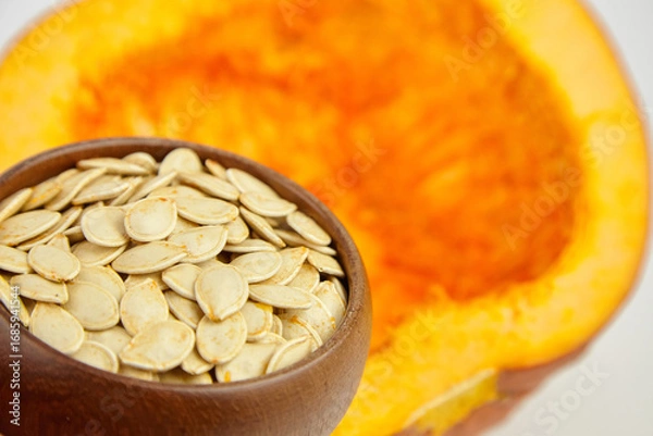 Fototapeta Close-up of a wooden bowl filled with pumpkin seeds, set against a vibrant orange pumpkin half, showcasing natural textures and colors in a seasonal context