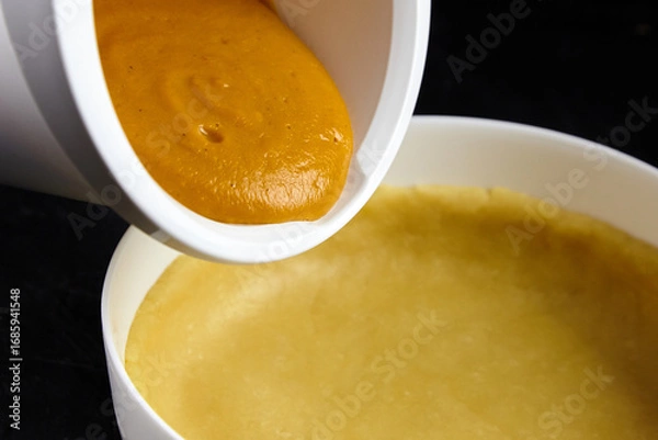 Fototapeta Pumpkin filling being poured from a white bowl into an unbaked pie crust, showcasing the preparation of a delicious dessert with rich textures and vibrant colors