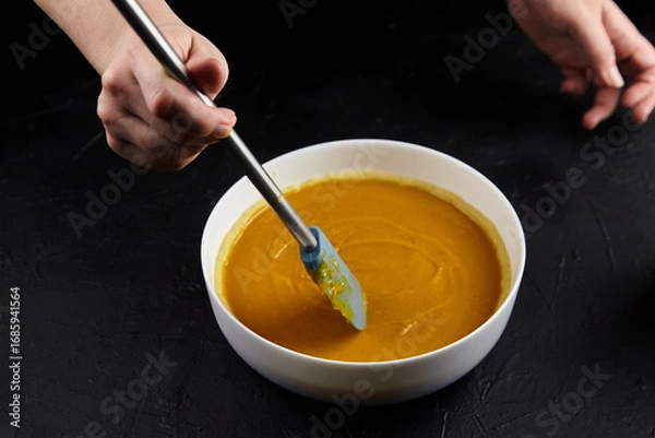 Fototapeta Hands of a person stirring creamy pumpkin puree in a white bowl with a spatula on a dark textured surface, showcasing culinary preparation and vibrant colors