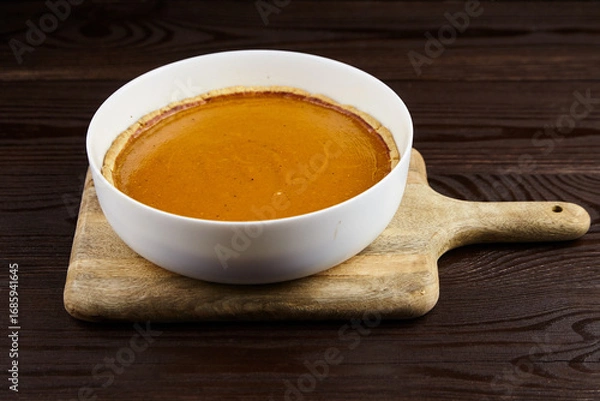 Fototapeta Creamy pumpkin pie in a white bowl placed on a wooden cutting board, showcasing rich texture and warm color, perfect for autumn dessert presentations