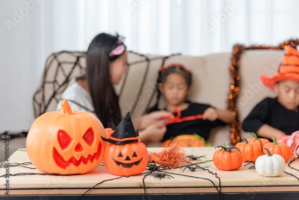 Fototapeta Pumpkins with smiling faces and witch hats decorate a table, with children in costumes visible in the background celebrating halloween day, black spiders and halloween decor also are present