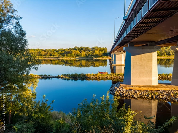 Obraz View from the Vistula riverbank under a large highway bridge, with people fishing from a red  boat.