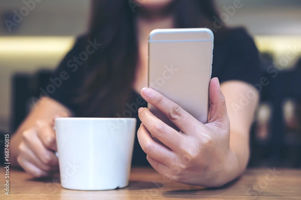 Fototapeta Closeup image of a beautiful Asian woman using and looking at smart phone while holding a white coffee cup in modern cafe