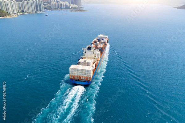 Fototapeta Big heavy container ship leaves the bay and strait for the open ocean near a metropolis with multi-story buildings